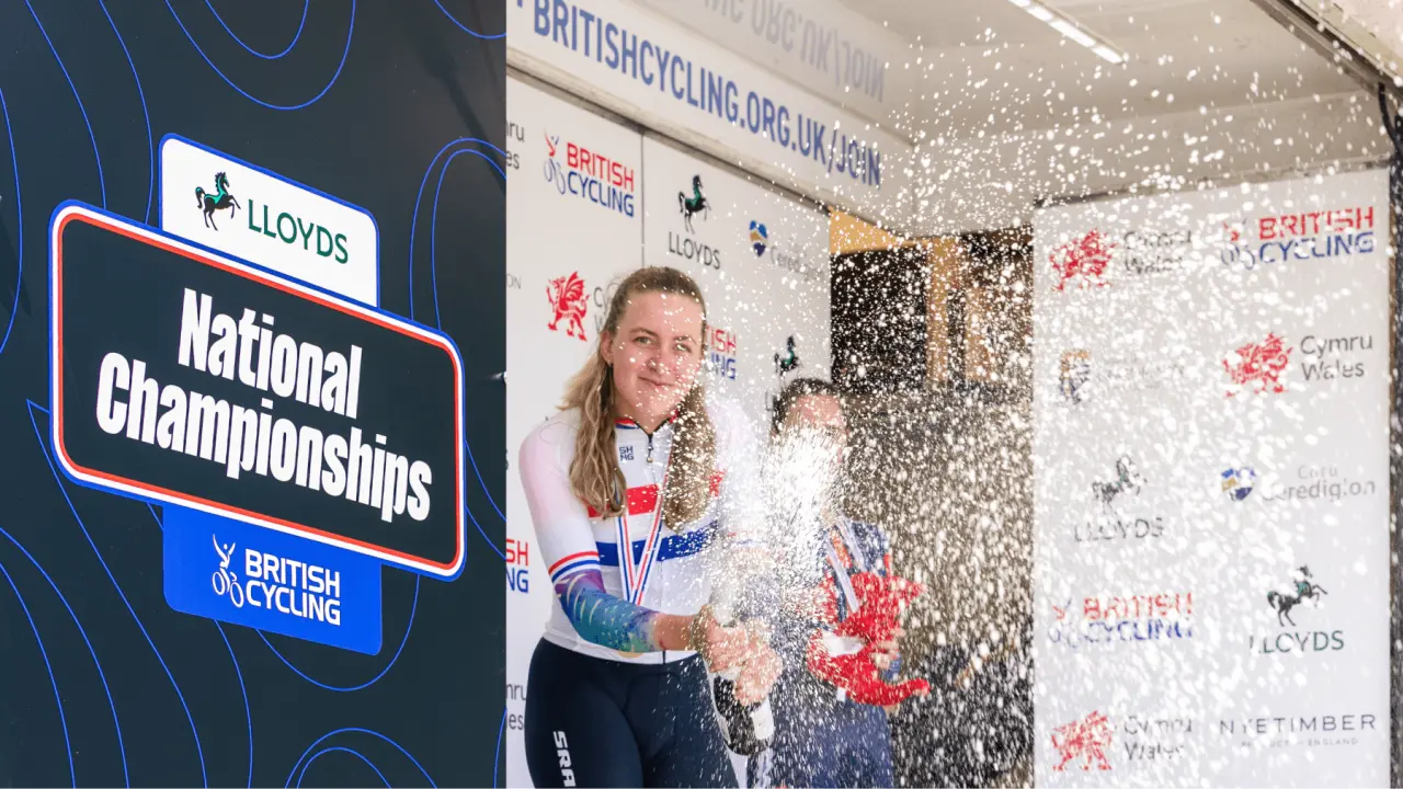 Cyclist spraying champagne on podium with Lloyds National Championships signage and British Cycling branding visible behind her
