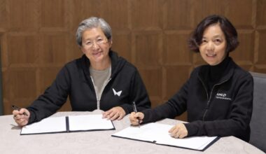 Two women in black hoodies—one from AMD wearing a World Labs hoodie, and one from World Labs wearing an AMD hoodie, smile at the camera while sitting a desk with papers to sign on it.