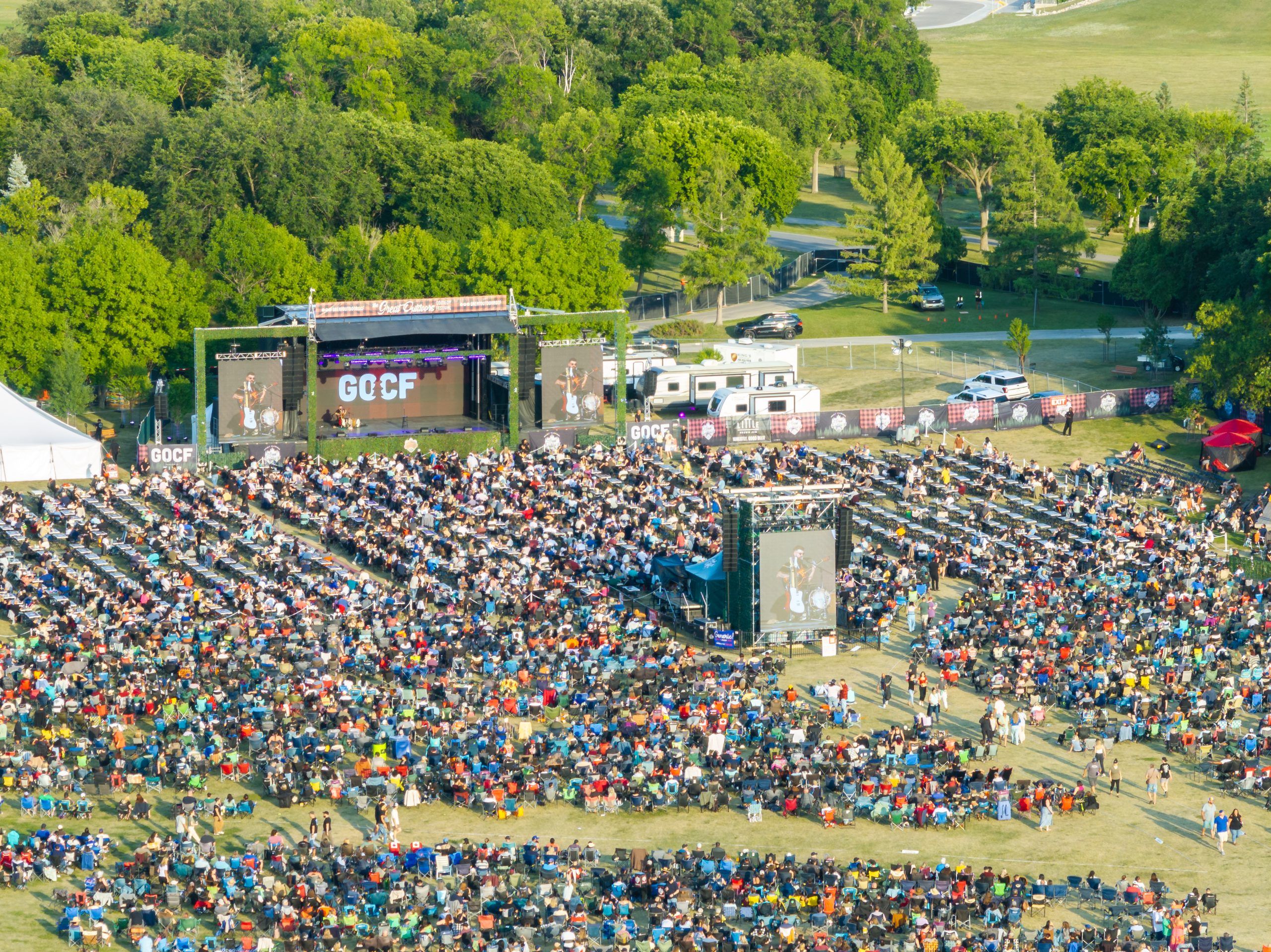Crowd of people in an outdoor setting within trees and near a stage.