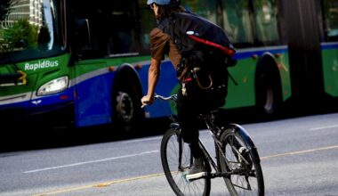 A man wearing a brown shirt, a backpack and a cap can be seen from behind as he rides a bicycle on a city street. A bus can be seen in the background.