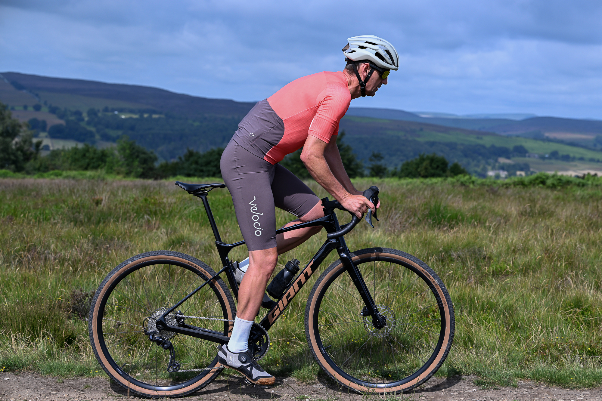 man riding a black gravel bike wearing brown shorts, orange top, white socks and grey shoes on a gravel trail