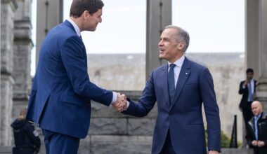 Two men in blue suits shake hands outdoors in front of a building with tall concrete columns.