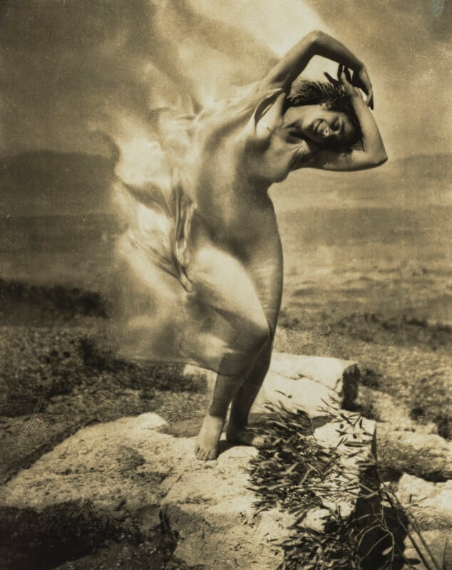 A woman with arms raised above her head dances on a stone platform, her sheer garment blurred by motion against a landscape.