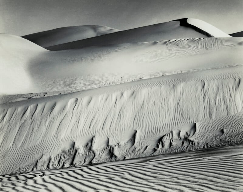 A black-and-white landscape of rolling sand dunes with prominent wind-swept ripples and deep shadows.