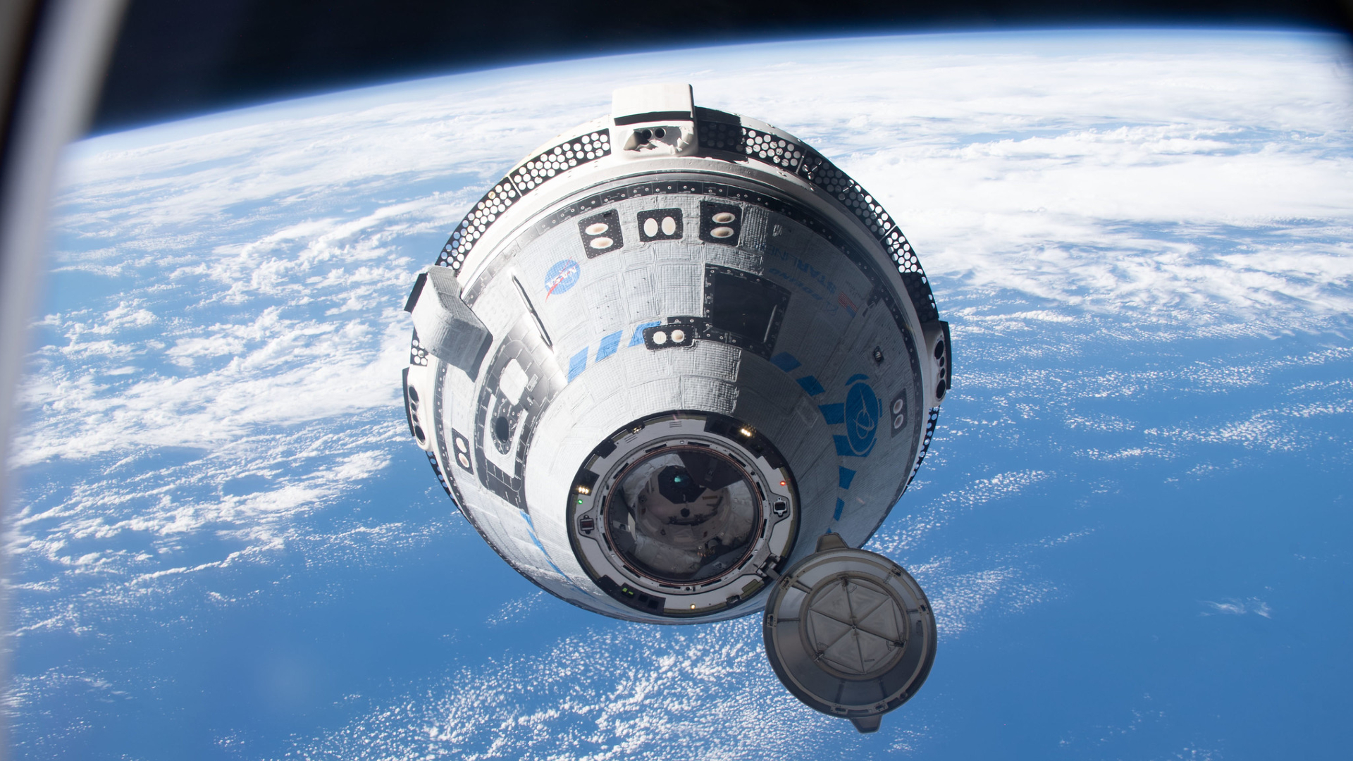 A white and blue Boeing Starliner capsule floats above a blue Earth with the black of space in the background.