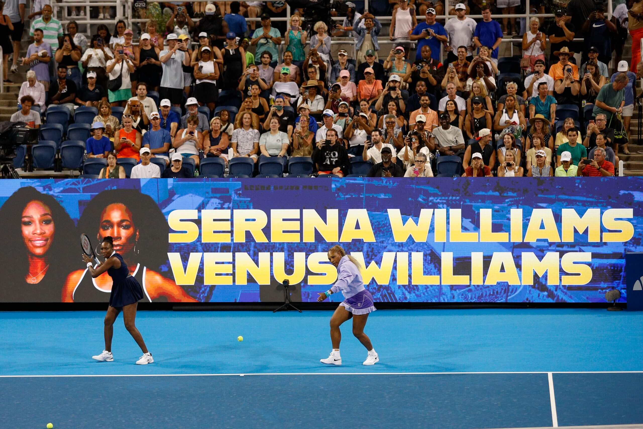 Serena and Venus Williams warm up on a blue tennis court with fans behind them.