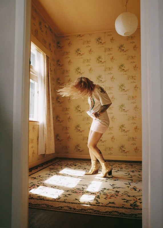 A person with long hair whipping in motion stands in a room with floral wallpaper, wearing a t-shirt, skirt, and gold boots.