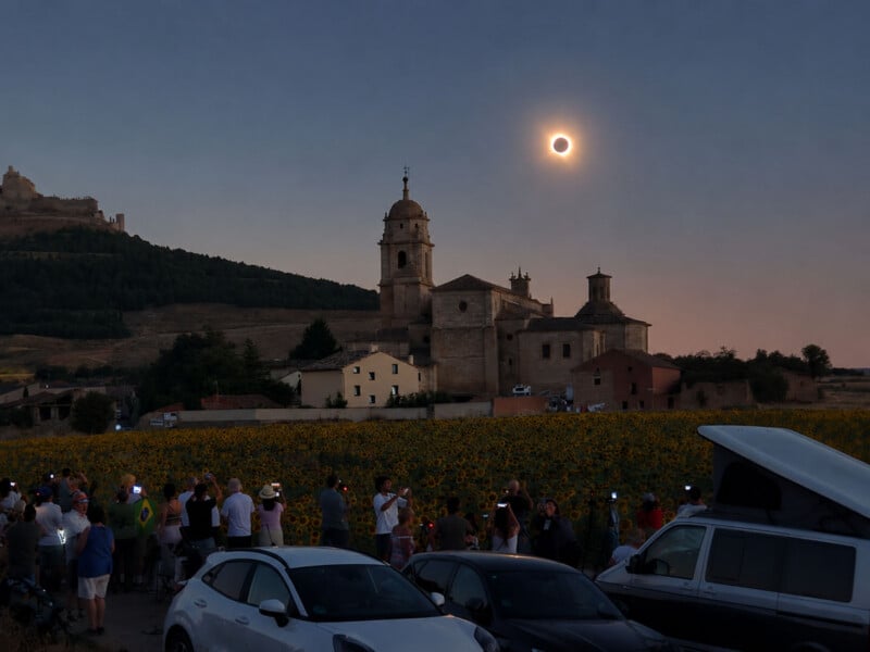 A crowd of people stands in a sunflower field at dusk, watching a solar eclipse above a stone church and a distant castle ruin.
