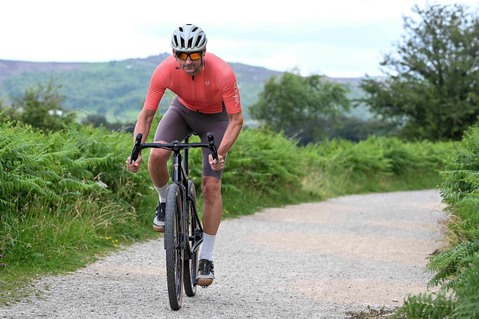 man riding a black gravel bike wearing brown shorts, orange top, white socks and grey shoes on a gravel trail towards the camera