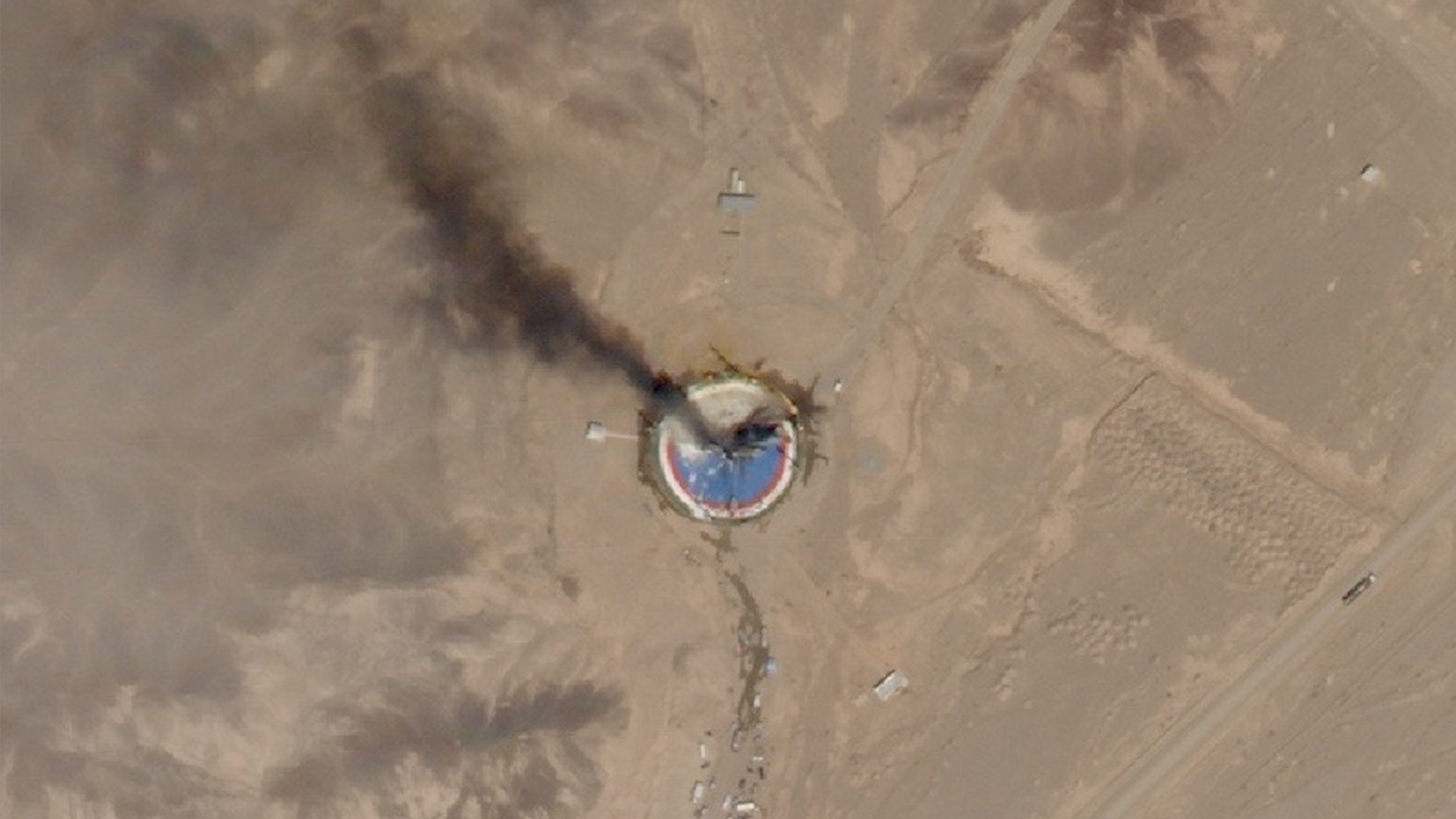 satellite photo of a dark plume of smoke ascending from a blue and red circular launch pad in a desert landscape