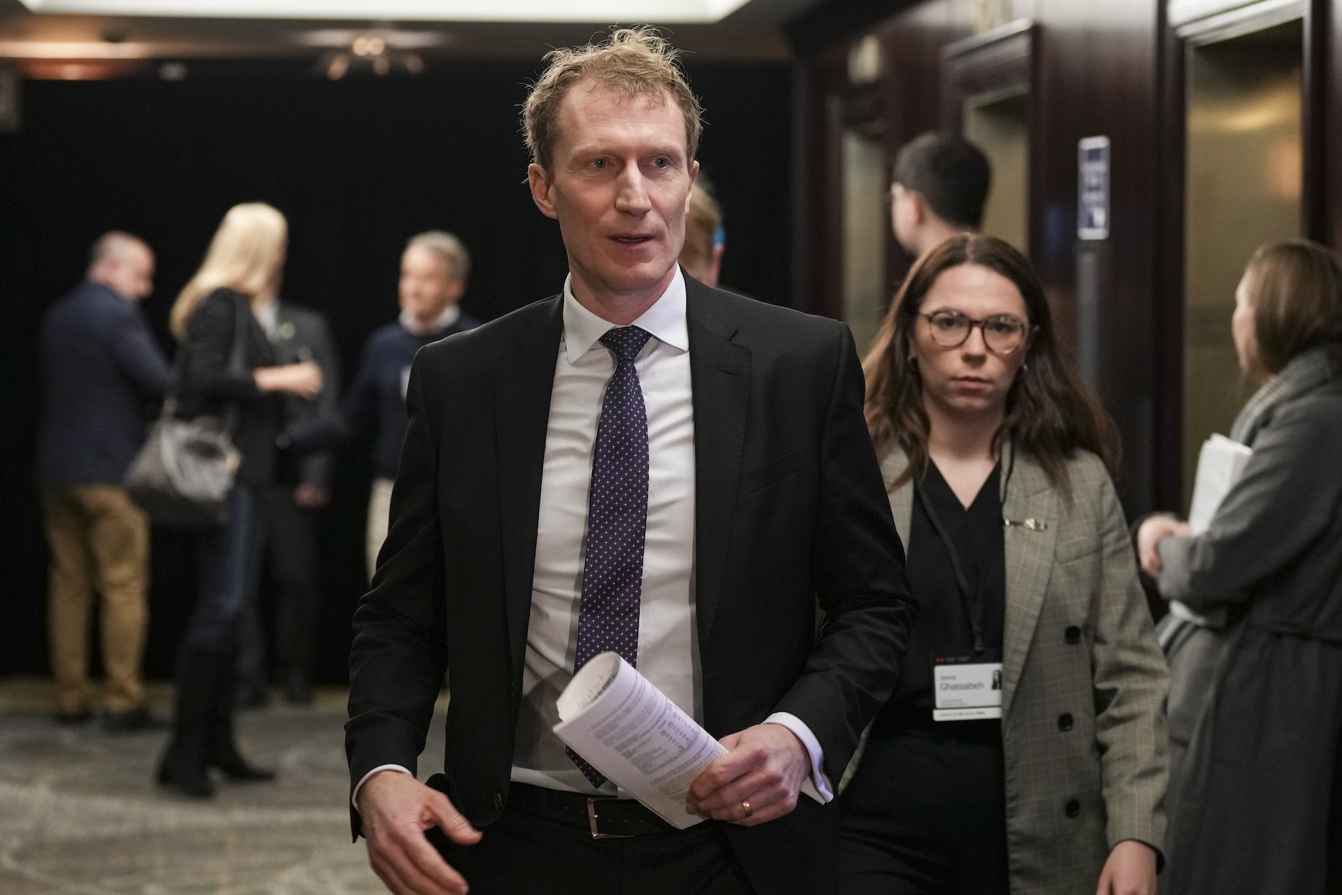 A head-on shot of Marc Miller walking past a bank of elevators with an aide behind him. He's carrying papers in his left hand.