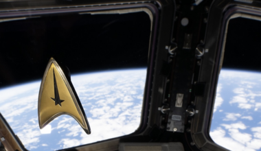 A golden Star Trek delta badge with a tall black star at its point floats in front of windows showing Earth from the ISS.