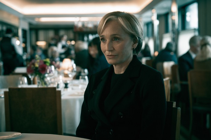 Kristin Scott Thomas in Slow Horses, wearing all black and sat at a table in a restaurant.