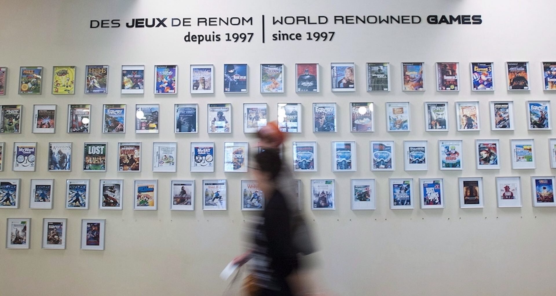 A shot of dozens framed video-game covers on a wall, with the label "World renowned games since 1997" inscribed above them. There are two passers-by blurred in the foreground.