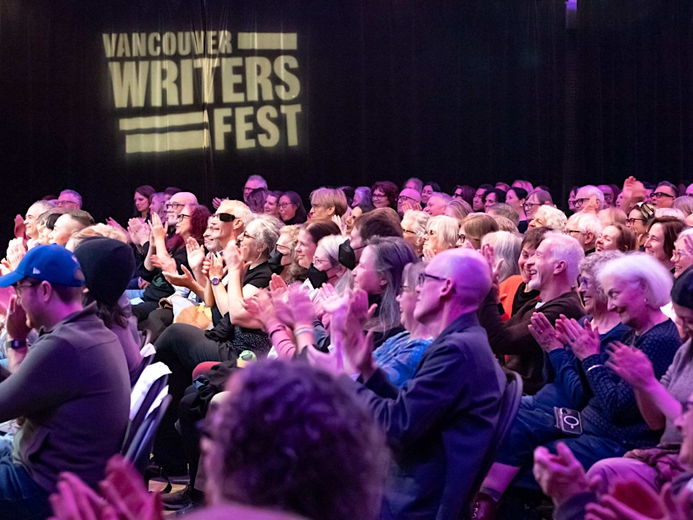 An audience applauds under purple lights with the words ‘Vancouver Writers Fest’ projected on a wall.