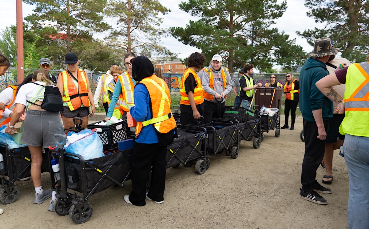 Around 10 people, many wearing high-visibility orange and yellow safety vests, stand near wheeled carts in an outdoor setting.