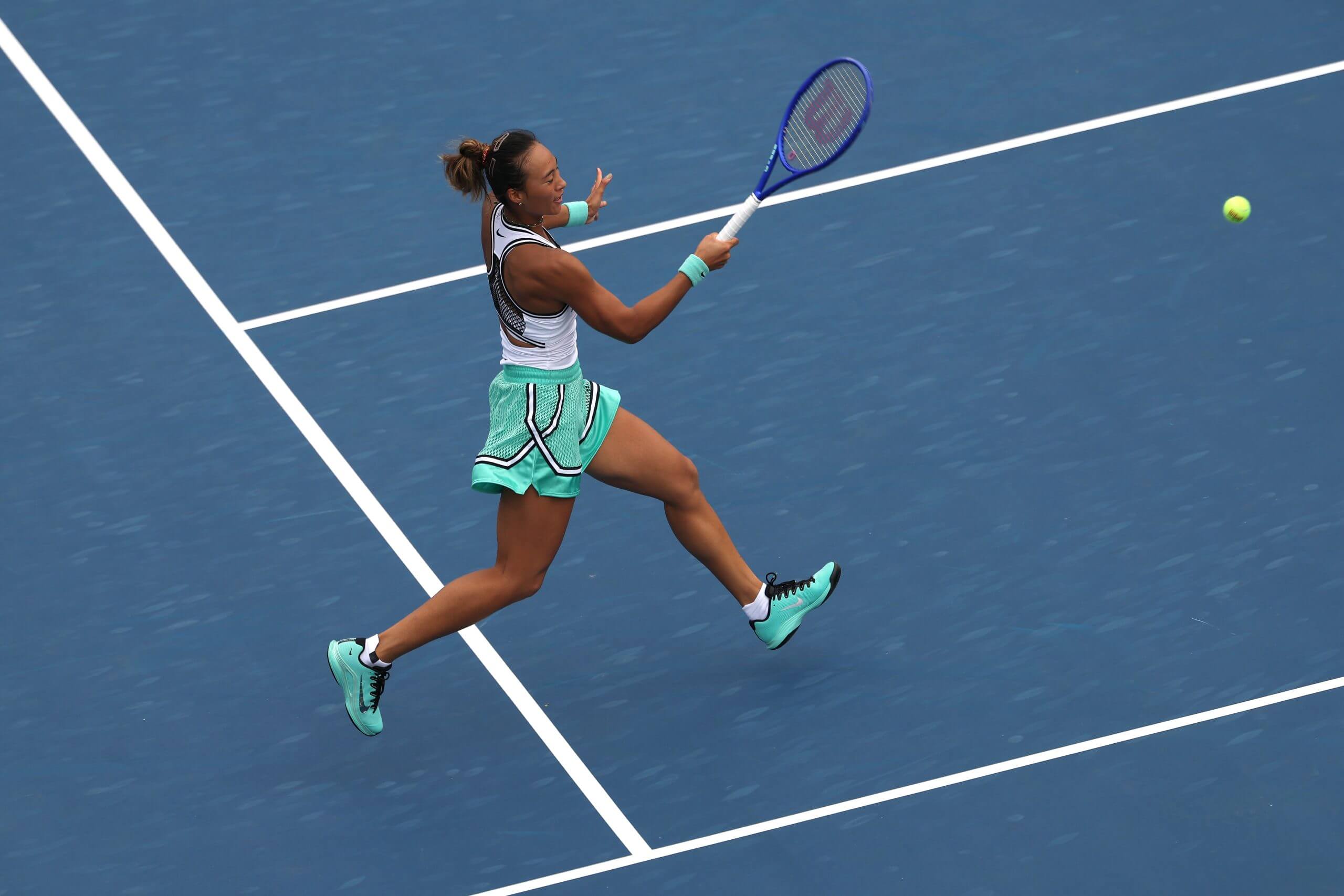 Zheng Qinwen hits a forehand on a blue tennis court.