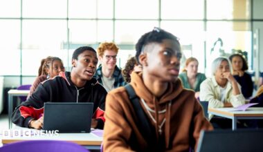 Students sit at desks with laptops in a bright classroom or lecture space. Several people are facing forward as if listening to a lesson, with large windows letting in daylight behind them.