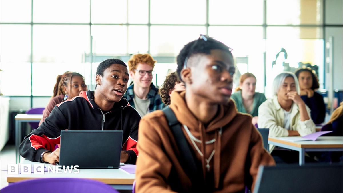 Students sit at desks with laptops in a bright classroom or lecture space. Several people are facing forward as if listening to a lesson, with large windows letting in daylight behind them.