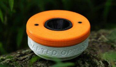 A 3D-printed, two-tone orange and white circular device with the words "ADAPTED OPTICS" embossed on the side, resting on moss.