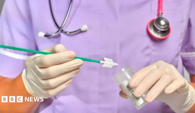 A medic, pictured from the shoulders to waist wearing purple scrubs, with a stethoscope around their neck, wearing white gloves and holding a sample tube.