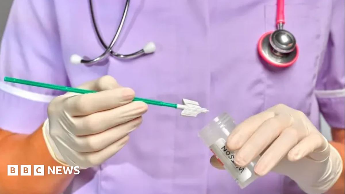 A medic, pictured from the shoulders to waist wearing purple scrubs, with a stethoscope around their neck, wearing white gloves and holding a sample tube.