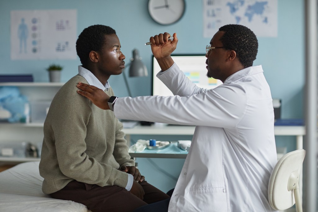A male doctor examines a young man's eyes with a penlight in a clinic.