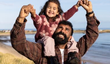 A waist-up shot of a father carrying his young daughter on his shoulders as they walk along the coast on a beautiful, sunny day. They are both smiling, enjoying their time together by the ocean