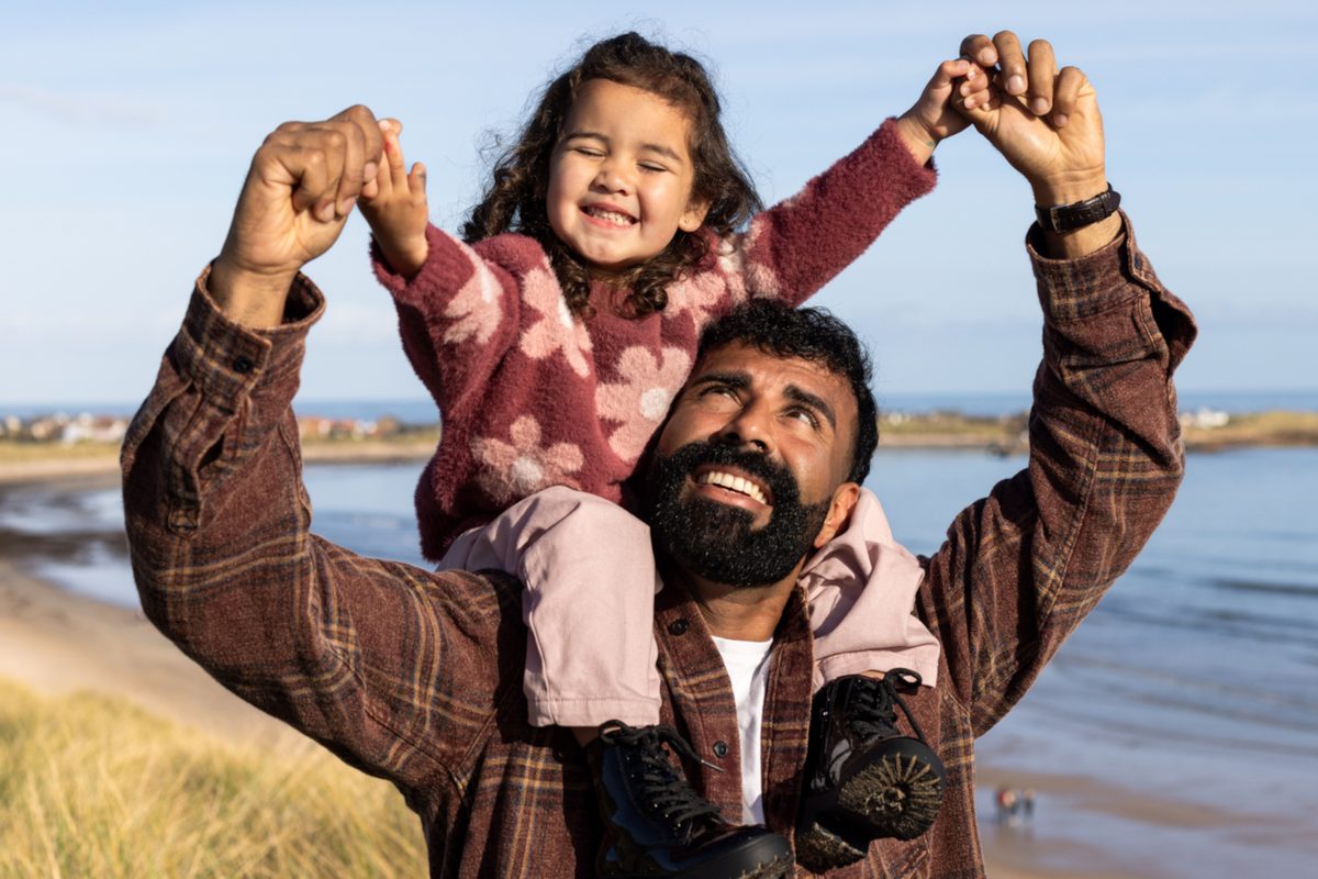 A waist-up shot of a father carrying his young daughter on his shoulders as they walk along the coast on a beautiful, sunny day. They are both smiling, enjoying their time together by the ocean