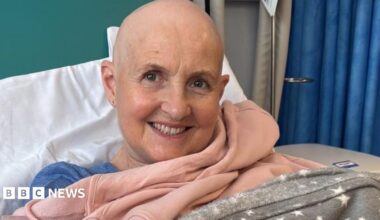 Caroline Ainslie in a hospital bed she is smiling at the camera. She has a bald head.