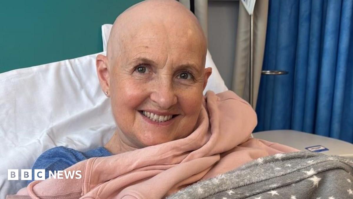 Caroline Ainslie in a hospital bed she is smiling at the camera. She has a bald head.