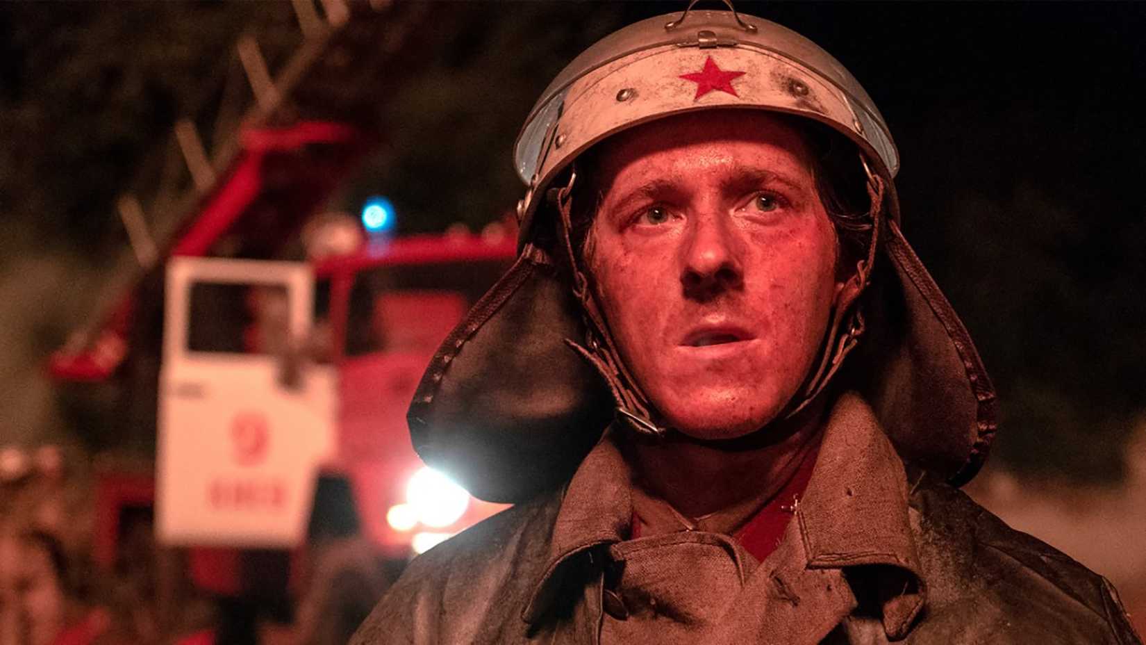 A firefighter with a red face staring out at something with a fire engine behind them in Chernobyl.