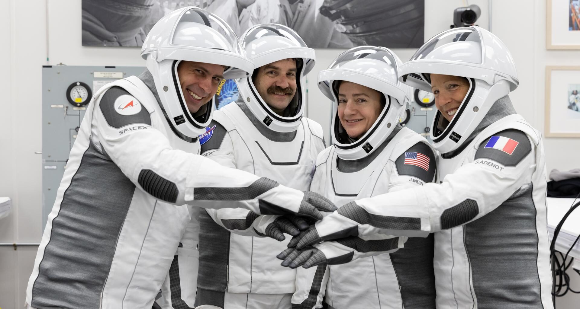 NASA’s SpaceX Crew-12 members stand side by side in their spacesuits with the face guard up. Each astronaut has their arm outstretched in front of them to pile their hands on top of one another as they smile and pose for a team photo. The astronauts are at the Neil A. Armstrong Operations and Checkout Building at the agency’s Kennedy Space Center in Florida ahead of launch to the International Space Station on Feb. 13, 2026, from left, Roscosmos cosmonaut Andrey Fedyaev, NASA astronauts Jack Hathaway and Jessica Meir, and ESA (European Space Agency) astronaut Sophie Adenot.