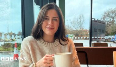 A close up image of Erin Atkinson wearing a cream jumper and holding a mug. She appears to be sitting in a public space with a car park seen out the window behind her.