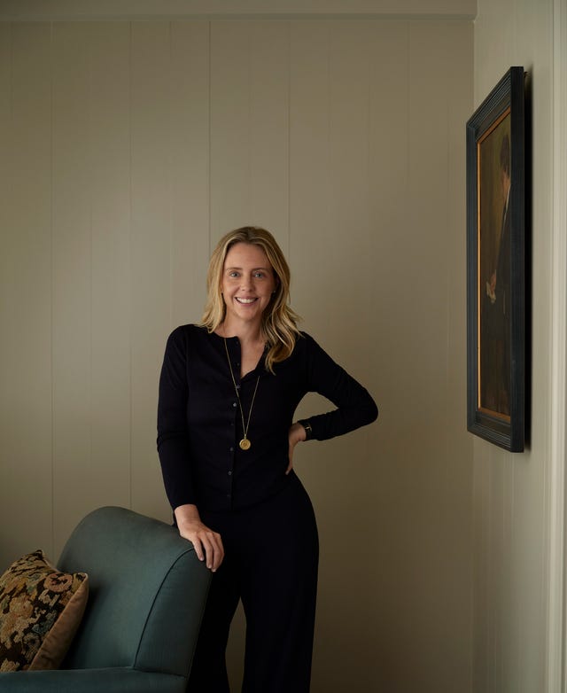 Smiling woman in a black outfit standing next to a green couch in a softly lit, minimalist room.