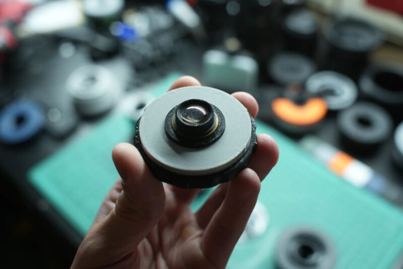 A hand holds a small, circular, 3D-printed camera lens assembly with a glass element in the center against a blurred workbench background.