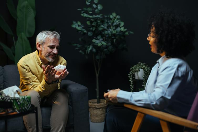 Older man sitting in an armchair across from a therapist, talking and holding a tissue