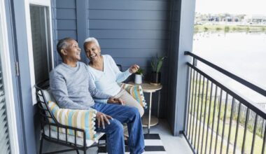 An older Black man and woman are sitting on a balcony, laughing and holding hands. The man, wearing a gray long-sleeved shirt and blue jeans, sits on the left. The woman, with short white hair and wearing a light blue long-sleeved top and khaki pants, sits on the right. They are on black metal patio chairs with striped cushions. Behind them is a dark gray siding wall and a sliding glass door. To the right, a black metal railing overlooks a calm body of water with buildings in the distance under a bright sky.