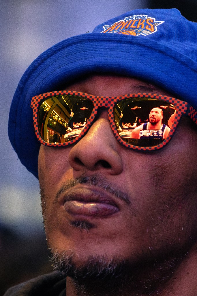 A fan wearing a New York Knicks hat and orange-and-black checkered sunglasses, with a reflection of Knicks guard Jalen Brunson in the lenses.
