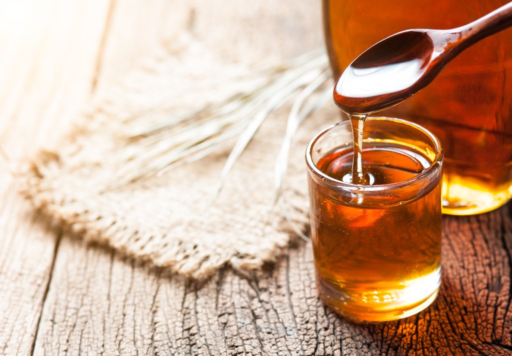 Maple syrup being poured from a spoon into a glass on a wooden table.