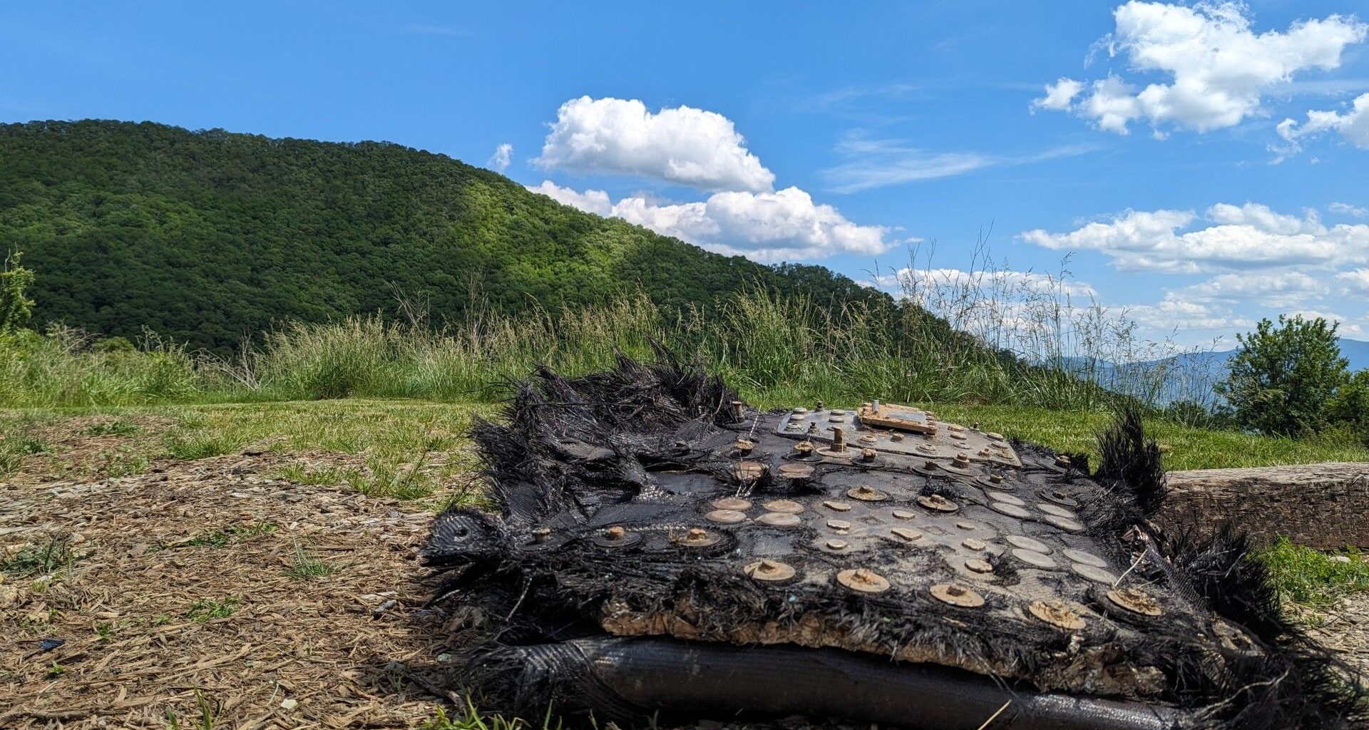 a large black piece of fiberglass covered in metal bolts and plates lies on the ground beside a trail leading into a forest. mountains can be seen rolling in the distance