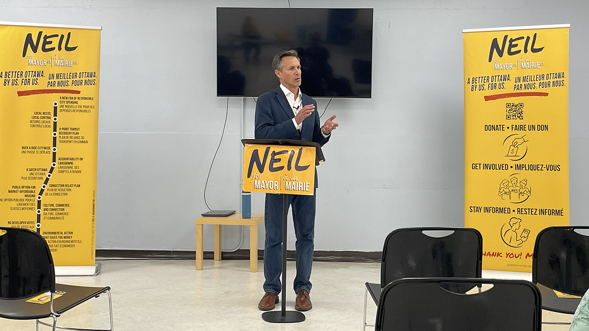 A man in a suit stands in front of a small podium before an audience. The room is decorated with yellow campaign signs reading "Neil for Mayor."