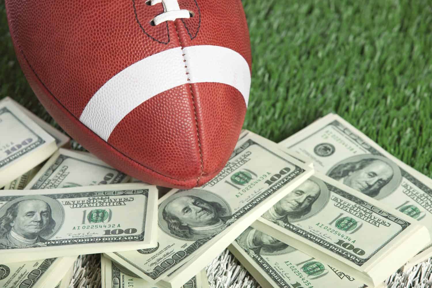 A brown American football with white stripes and laces rests on several scattered stacks of US one-hundred dollar bills. The money and football are positioned on a green surface resembling artificial grass or turf. The football is centered towards the top left, with stacks of money spread across the bottom and right of the frame.