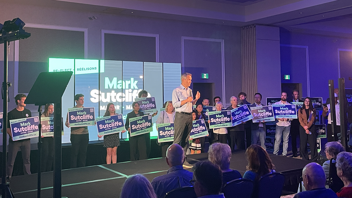 A man in a white dress shirt and black pants stands on stage with a microphone. Volunteers behind him hold signs reading "Mark Sutcliffe" and an audience sits in folding chairs in front of him.