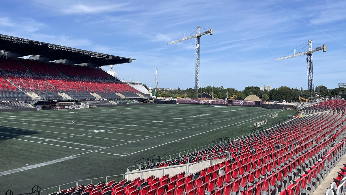 The field and stands of TD Place arena. In the background, there are construction vehicles and two large cranes in the skyline.