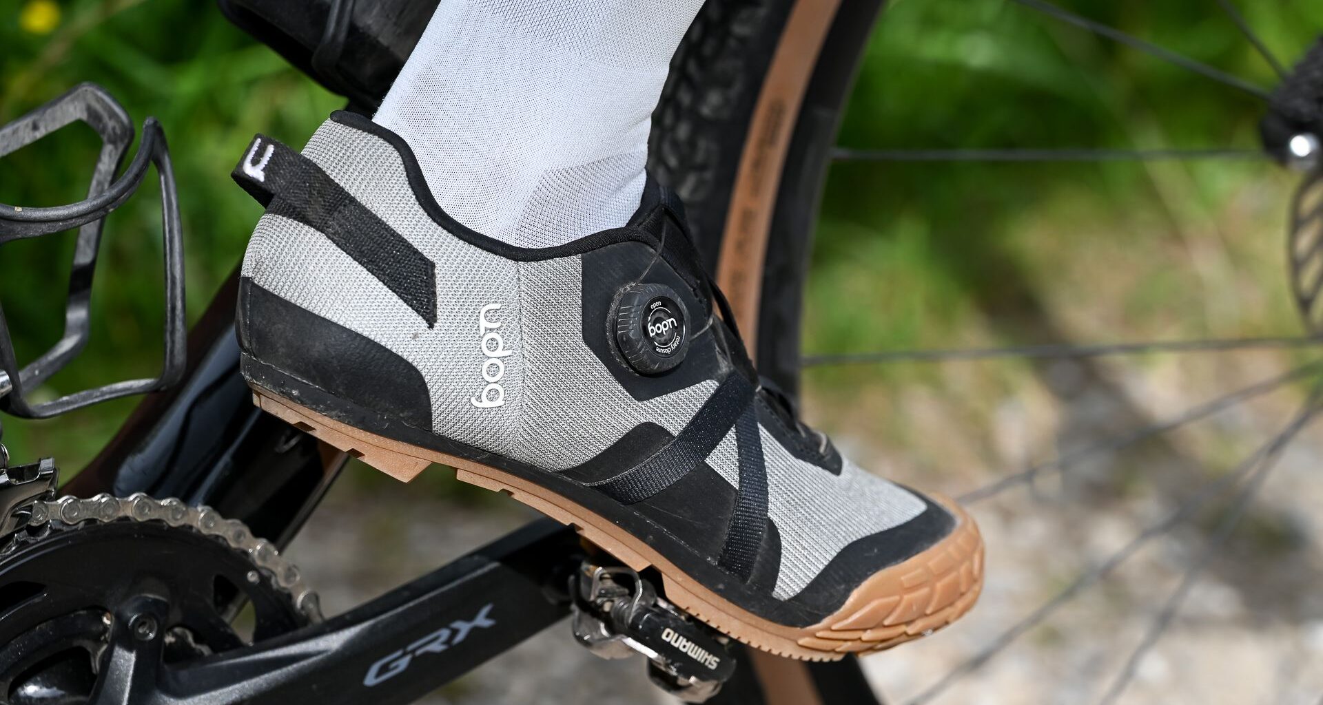 Close up of a foot wearing white socks and grey shoes clipped into a pedal on a black gravel bike