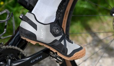 Close up of a foot wearing white socks and grey shoes clipped into a pedal on a black gravel bike