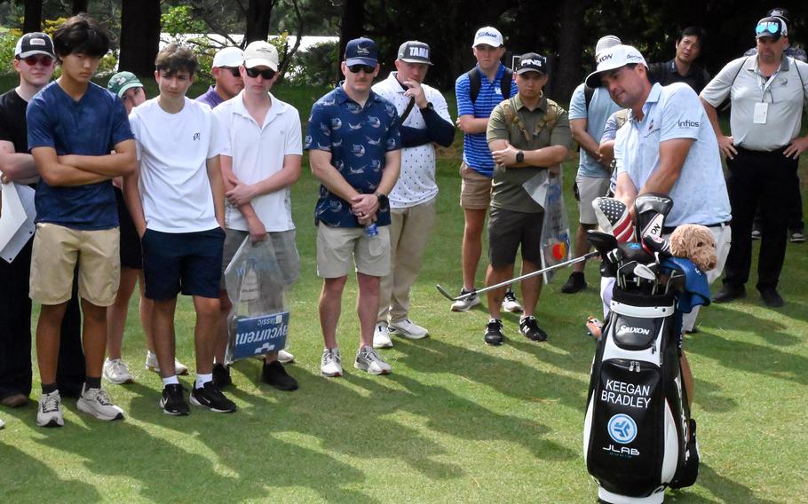 A professional golfer demonstrates a shot while a group of spectators gathers in a semicircle to observe. A golf bag filled with clubs stands in the foreground on the practice area.