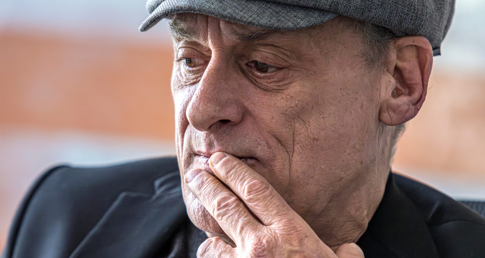An older man wearing a cap holds his fingers to his lips.