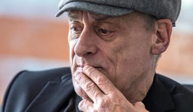An older man wearing a cap holds his fingers to his lips.